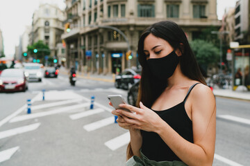 Young stylish woman in a black mask ordering a taxi by a mobile app on a smartphone to go home avoiding public transport. Woman wearing protective mask while standing on a street with a mobile phone.