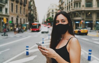 Portrait of a brunette woman wearing mask waiting for a taxi she ordered by the mobile phone application. Young woman in a mask looking aside while standing on a crossroad with a mobile phone in hands