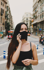 Young girl wearing black sterile medical face mask talking by the mobile phone while standing on a street. Attractive woman in a mask calling to the hospital to check the coronavirus test results
