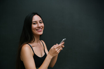 Young brunette caucasian woman with long hair texting online by a smartphone while standing on a black street wall background. Stylish girl smiling at the camera while holding a mobile phone in hands.
