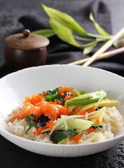 Japanese cuisine. Rice with avocado, salmon, vegetables, caviar and herbs in a deep white plate on a black table. Restaurant menu. Background image, copy space
