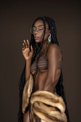 A portrait of a serene young black female with long dreadlocks, beautiful makeup and moist lips posing by herself in a studio with dark background wearing jewelry, a bikini & fur coat.