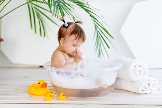 little baby girl washes and plays in a basin with foam and water in a bright room at home