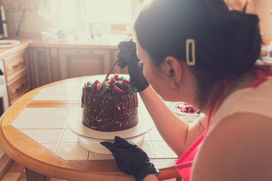 Confectioner young woman decorates with a brush and food coloring chocolate cake decoratedwith strawberries, blueberries and a pomegranate.