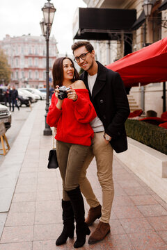 Full height outdoor image of fashionable elegant couple in love walking on the street during date or holidays.  Brunette woman in red sweater making photos by camera.