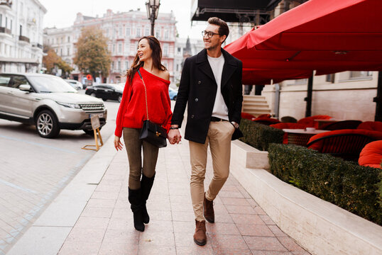 Outdoor fashion image of elegant couple walking in city. Happy romantic moments. Lovely girl in red sweater looking on handsome man in stylish jacket. European holidays.