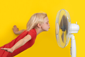 Cute boy refreshing from the heat in front of the fan. Hot weather. Child and fan on a yellow...