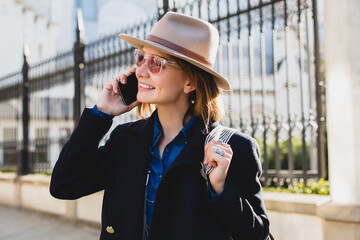 Fototapeta premium young stylish pretty woman smiling, happy, surprised, talking on her phone, dressed in dark blue coat and denim shirt, pink sunglasses, with a hat and backpack, sunny day, good weather, city street