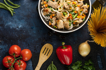 Top view of pan with frying chicken meat with vegetables for pasta on dark blue background. Professional kitchen table with wooden spatula, tomatoes, red pepper, onion and other ingredients for dish.