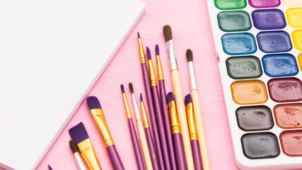 Naklejka premium Watercolor paints with brushes and an album on a pink background.
