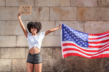 Black woman with afro hair holds the American flag and a sign with the message Black Lives Matter in the sunlight. Fight against racism.