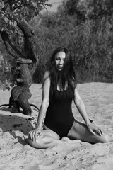 Slim long-haired young woman in bikini sitting on the sand near old tree in black and white