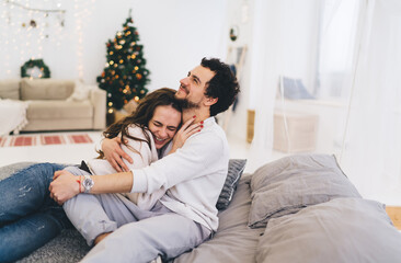 Funny caucasian male and female lovers enjoying free time and winter vacation celebrating at home interior, couple in love sitting at festive decorated apartment hugging and feeling happiness together