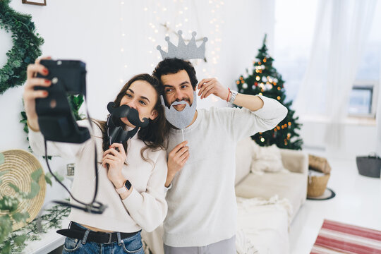 Funny young caucasian couple in love having positive photos using masks celebrating new year at apartments, girlfriend using decoration with boyfriend for taking photos on camera at festive flat
