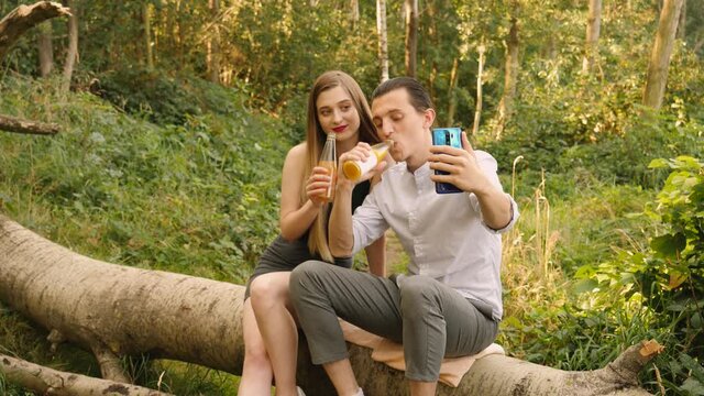 Young people couple date spend time together using phone take selfie pictures for social media drink bottled sweet beverage sitting on fallen tree trunk on nature countryside