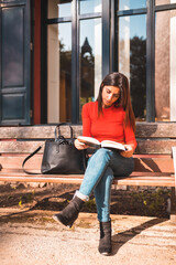 Lifestyle. Young Caucasian brunette in a red shirt sitting on a bench in the park reading a book. Vertical photo