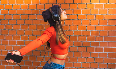 Lifestyle, young brunette with a wool cap and a red sweater with her eyes closed listening to music from her cell phone with her black headphones. Background of a brick wall