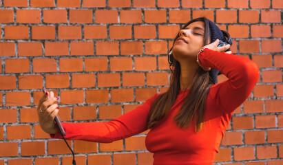 Lifestyle, young brunette with a wool cap and a red sweater enjoying music with her black headphones. Background of a brick wall