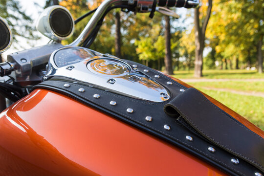 Close up photo of a tank of orange motorcycle with lots of chrome details, music speakers and riveted tie, made of genuine brown leather. Motorcycle is shot in the autumn park against yellow trees