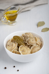 Homemade pelmeni or dumplings of Russian cuisine made of minced meat filling wrapped in dough served in white bowl with black pepper seasoning and bay leaf on white wooden background. Vertical