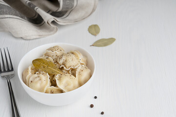 Pelmeni or Russian dumplings made of minced meat filling wrapped in dough served in white bowl with fork with black pepper seasoning and bay leaf on white wooden background. Image with copy space