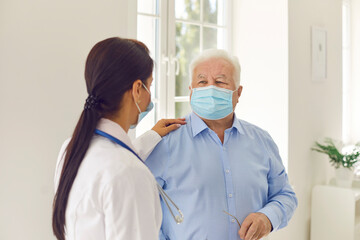 Senior patient and young female doctor, wearing medical face masks, communicating in hospital office
