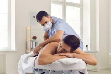 Young man enjoying body massage done by professional masseur or physiotherapist in medical face mask