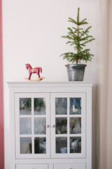 Christmas tree in a metal bucket, vintage wooden red toy horse on a white dresser in the interior of the living room, decorated for Christmas and New Year