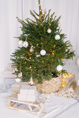 Christmas decorated tree in a wicker pot decorated with white toys. Next to the tree there are wooden decorative sleds with gifts