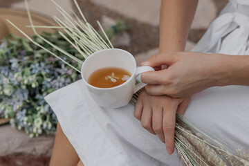 Cup of tea in female hands of a young girl outdoor, with blurred flowers and summer hat on background. Young woman in white dress is holding spikelets and mug with tea and flowers inside. .