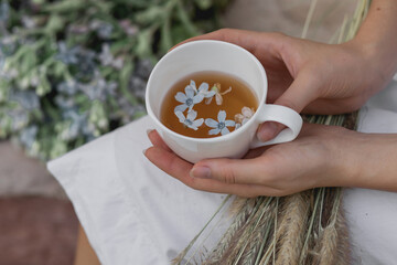 Cup of tea in female hands of a young girl outdoor, with blurred flowers on background. Mug with tea and blue flowers inside. .