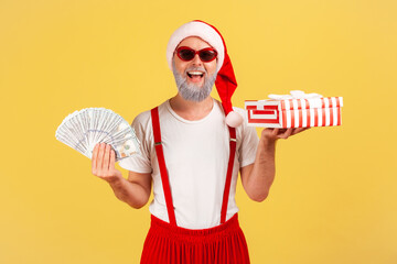 Cheerful positive elderly man in sunglasses and santa claus hat holding fan of dollars and gift box smiling at camera, preparing presents on holidays. Indoor studio shot isolated on yellow background