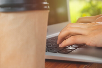 Close-up of hands using laptop on the wooden table. Hands typing on the keyboard. Blurred of green leaf background. Working out door concept.