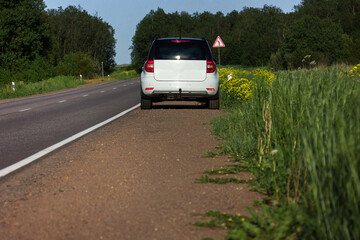 Fototapeta na wymiar A modern white car sits at the edge of the road next to a green field while traveling and looking for new routes. Outdoor weekends, local travel and summer trips