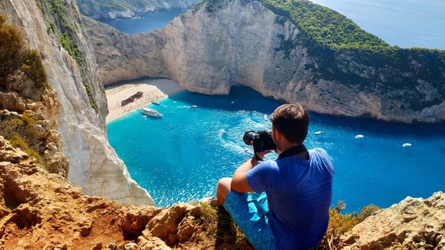 A man photographer with DSLR camera sits on top of a mountain on a cliff overlooking the sea and navayo beach on the island of Zakynthos in Greece