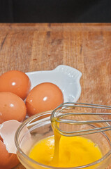 Top view, medium distance of a raw, scrambles egg in glass bowel with whisk, and egg shells in ceramic, white bowel, on a wood cutting board and black background