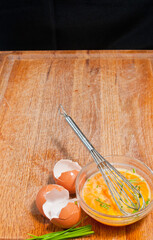 Top view, medium distance of a raw, scrambles egg in glass bowel with whisk, egg shells with, and three brown eggs in white bowel, cut chives on a wood cutting board and black background