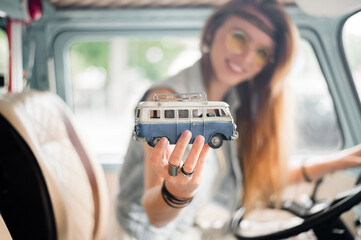Young hippie woman driving a classic van with a toy car model in her hand. Smiling blurred girl...