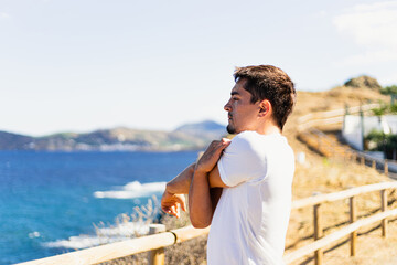 Young sportsman stretching arms outdoors while looking at the horizon with the sea to the side.