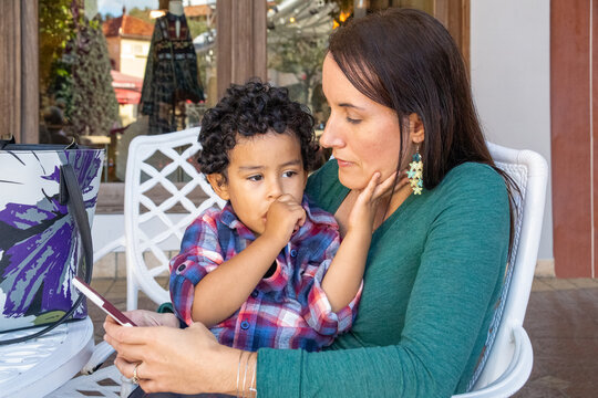 A young mother sits with her toddler boy to comforts and soothes outside the store, texting on her smartphone. The young boy is relaxed in his mother's arms, sucking his thumb while touching mommy.