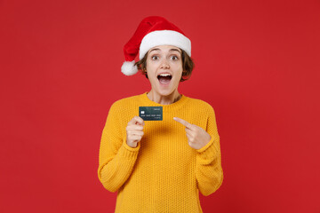 Shocked young Santa woman in casual yellow sweater Christmas hat pointing index finger on credit...