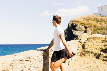 Young sportsman stretching his legs outdoors while looking at the horizon with the sea to the side.
