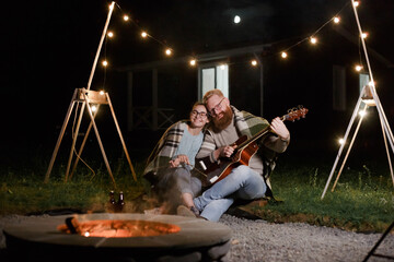 Fototapeta na wymiar Young couple, caucasian woman and red haired bearded man, on a night picnic with a guitar and marshmello have a good time