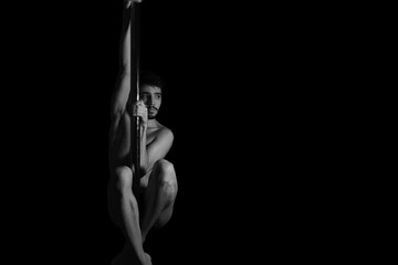 Dramatic dark portrait of a young man hanging from a pole dance bar with his eyes opened. Dark background studio shot. Black and white image.