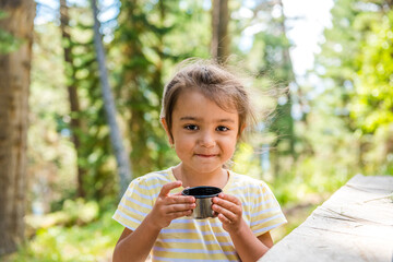 Cute little girl in a summer forest holding a cup of tea and smiling. walk to drink hot tea from a thermos. Seasonal outdoor activities