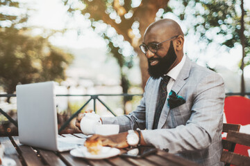 A cheerful handsome bald black businessman in eyeglasses and neat beard is having a coffee break in an outdoor restaurant, pouring himself some tea while reading a chat message on his laptop in front - Powered by Adobe