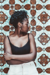 A vertical shot of a lovely young black female in a tank top and eyeglasses, with a curly Afro hair, standing in front of a tiled patterned Azulejo wall traditional for Portugal and looking aside