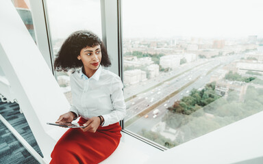 A beautiful African-American woman entrepreneur in a white blouse and red skirt is holding a digital tablet pc while sitting indoors on the bench on the top floor of a business office skyscraper - Powered by Adobe