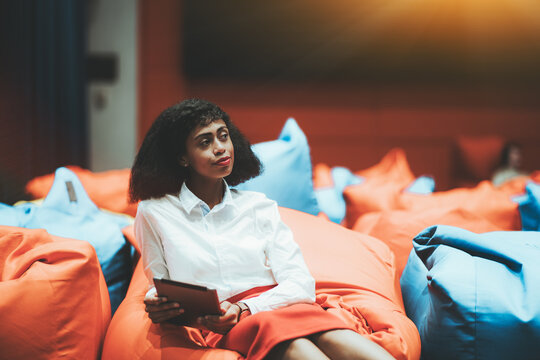 Portrait of a young beautiful African-American businesswoman sitting on an orange cushion with a tablet pc in hands indoors of a modern office chillout coworking area and thoughtfully looking aside