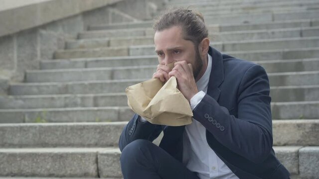 Stressed nervous Caucasian man breathing into paper bag. Portrait of depressed businessman sitting on urban city stairs having panic attack. Overworking problems.